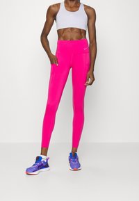 Aansluitende roze legging gemaakt van rekbaar materiaal met zijzakken, gecombineerd met een lichtgrijze sportbeha en blauwe sneakers.