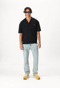 Chemise noire à manches courtes avec deux poches, jeans bleu clair et baskets jaunes et blanches, se tenant devant un fond uni.
