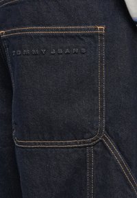 Jeans din denim albastru închis, cu cusături portocalii. Include un buzunar din spate cu text embosat care citește "TOMMY HILFIGER." Design curat, greutate medie.