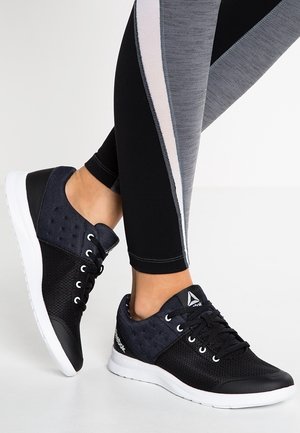 Pieds chaussés de baskets Reebok noires et grises avec semelles blanches, assorties à des leggings noirs et gris avec une bande blanche.