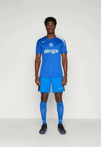 Man standing in blue Chelsea football kit with BingX logo, blue shorts, long socks, and black cleats against plain background.