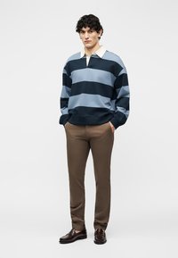 Νεαρός με σγουρά μαλλιά, φορώντας μπλε ναυτικό και απαλά γαλάζια ριγέ ριγέ ρούχα ράγκμπι, καφέ παντελόνι και σκουρόχρωμα καφέ μοκασίνια, στέκεται με τα χέρια στις τσέπες.