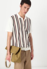 Kratka majica s kratkimi rokavi iz pletenine z navpičnimi črtami v kremni, rjavi in bež barvi. V kombinaciji z zeleno platneno torbo, ki jo drži model.