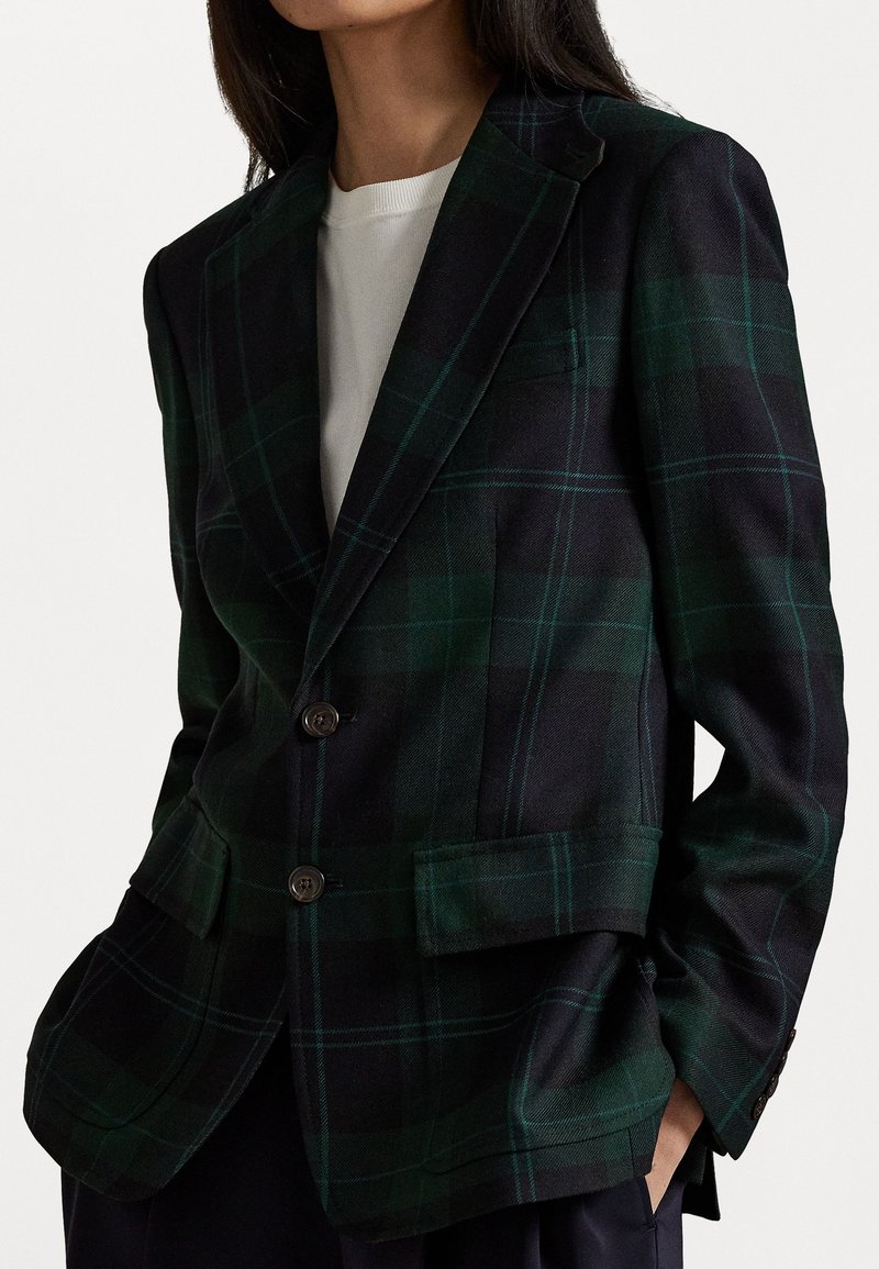 Donna che indossa un blazer a quadri verde scuro e blu navy sopra una maglietta bianca, con le mani nelle tasche, vista dalle spalle ai fianchi su uno sfondo semplice.