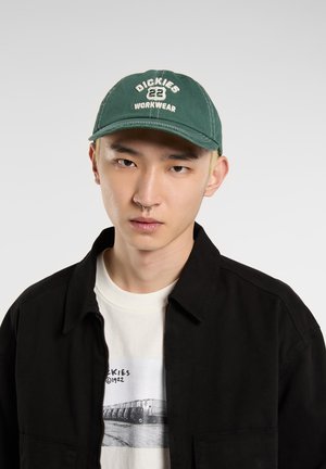 Casquette verte avec le texte brodé "Dickies 22 WORKWEAR", visière incurvée, en coton ; portée avec une veste noire sur un t-shirt graphique blanc.