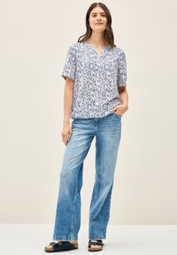 Blusa a maniche corte bianca con un motivo a foglia blu, design a scollo a V, abbinata a jeans a gamba larga blu chiaro e sandali scuri.