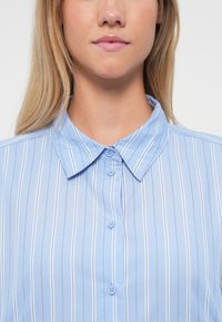 Nainen, jolla on päällään vaaleansininen paita, jossa on valkoiset pystysuorat raidat ja napillinen kaulus, näyttää alaosan kasvoista ja kaulasta.