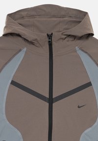 Veste à capuche de couleur marron clair avec des accents gris. Présente une fermeture éclair noire et un petit logo Nike noir. Tissu lisse avec des coutures contrastantes.