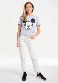 Graues Kurzarm-T-Shirt mit einem großen schwarz-weißen Mickey Mouse Grafik, kombiniert mit weißen schmal geschnittenen Hosen und schwarzen Turnschuhen.