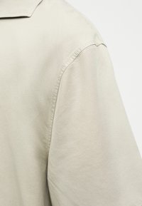 Κοντινό σε beige υφασμάτινο πουκάμισο που δείχνει λεπτομέρειες ραφής και άκρη του γιακά σε απλό φόντο.