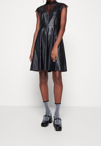 Schwarzes Lederkleid mit tiefem Spitzen-Ausschnitt, ärmellosem Design und fließendem Rock. Kombiniert mit silbernen Glitzer-Socken und schwarzen Blockabsatzschuhen.