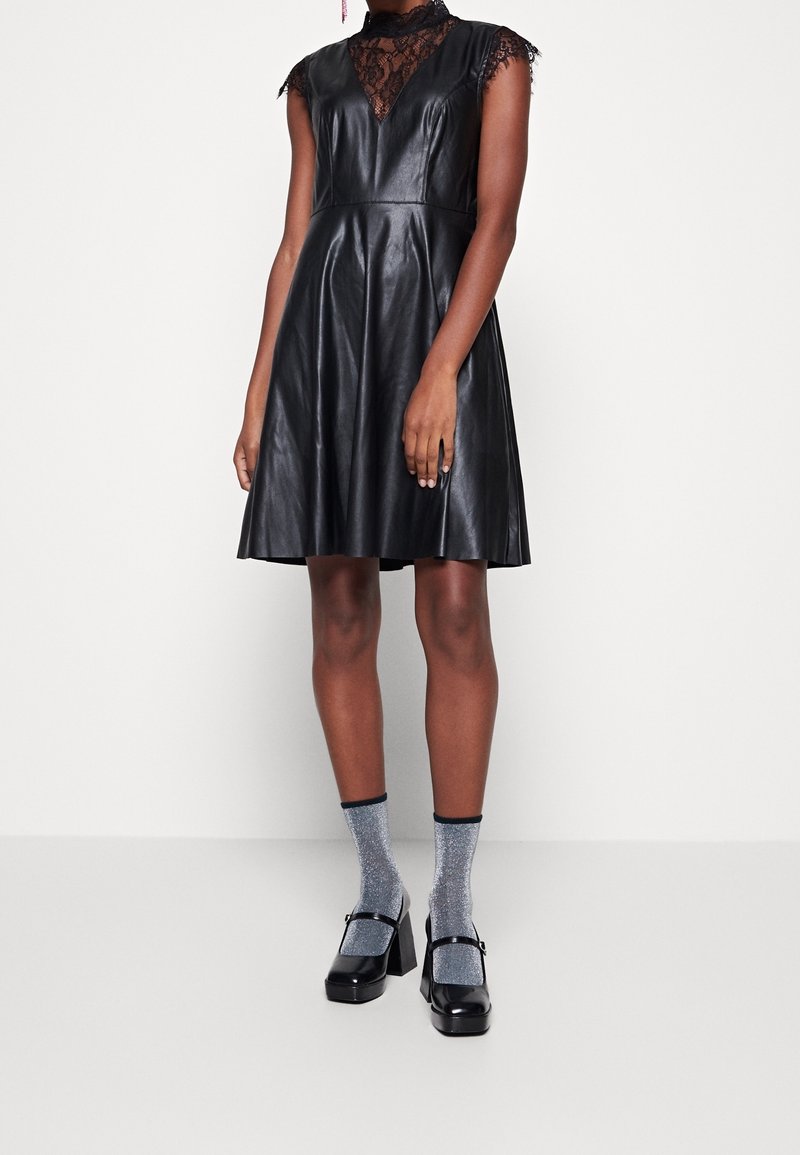 Schwarzes Lederkleid mit tiefem Spitzen-Ausschnitt, ärmellosem Design und fließendem Rock. Kombiniert mit silbernen Glitzer-Socken und schwarzen Blockabsatzschuhen.