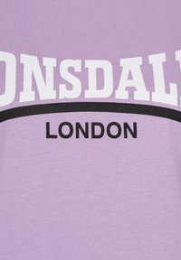 Svetlofialová bavlnená tričko s odvážnym bielym nápisom "LONSDALE" s čiernou zakrivenou čiarou nad ním a "LONDON" v čiernej farbe pod ním.