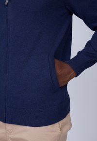 Veste zippée en tricot marine avec des détails en cuir brun sur les poches. Comprend des poignets et un ourlet côtelés, mettant en valeur une texture lisse et un design ajusté.