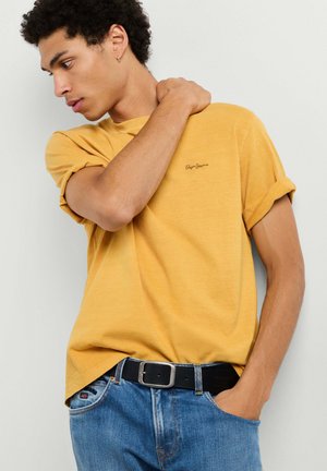 Junger Mann trägt ein gelbes T-Shirt mit hochgekrempelten Ärmeln und blaue Jeans mit schwarzem Gürtel, hält sich eine Schulter und schaut zur Seite.