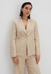 Blazer beige su misura con chiusura a un bottone, due tasche frontali, revers a punta e una texture liscia.
