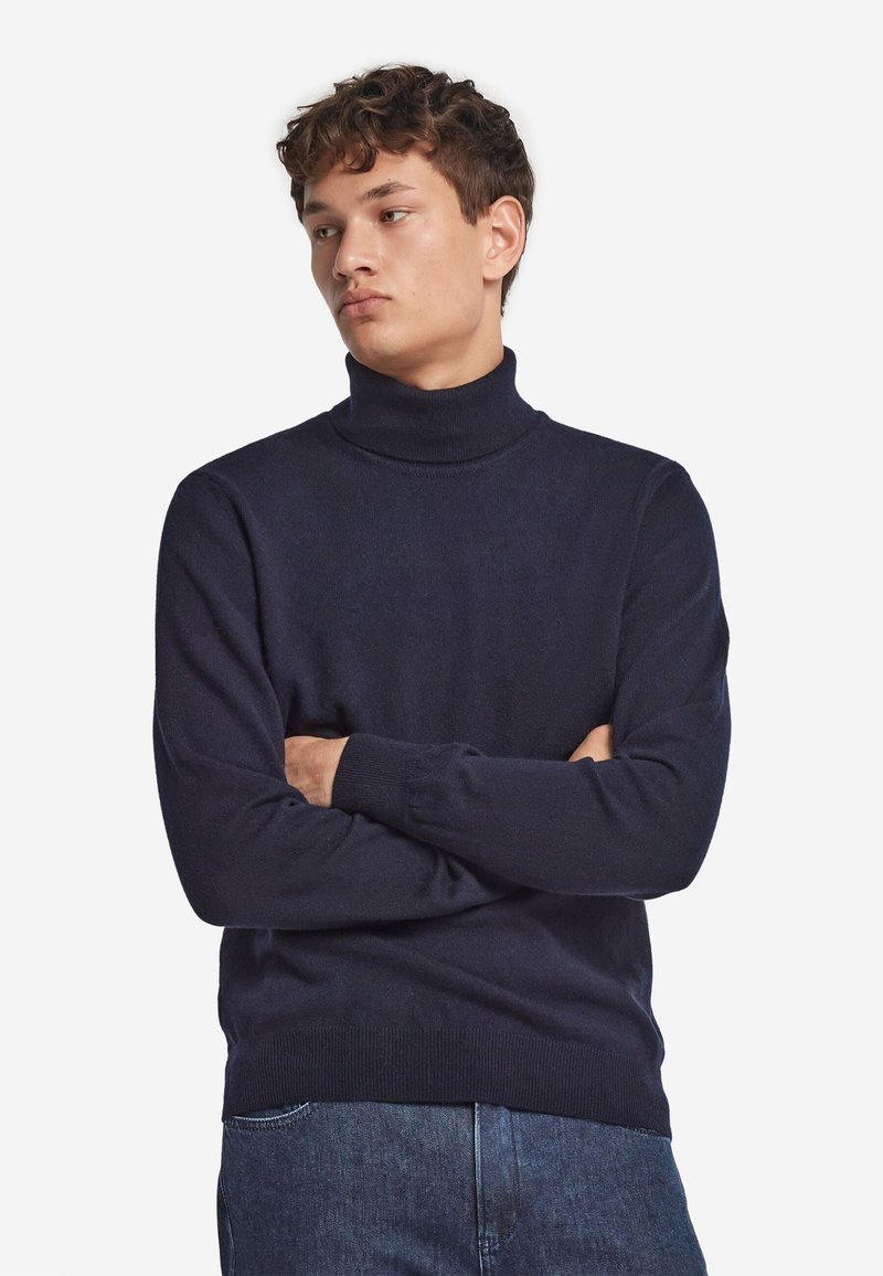 Junger Mann mit lockigem Haar, der einen dunkelblauen Rollkragenpullover und Jeans trägt, schaut seitlich mit verschränkten Armen.