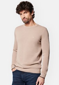 Jasno beżowy sweter z fakturą, z okrągłym dekoltem, d ługimi rękawami i ściągaczami; na piersi ma małe logo. Noszony z ciemnymi dżinsami.