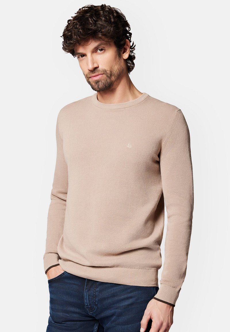 Jasno beżowy sweter z fakturą, z okrągłym dekoltem, długimi rękawami i ściągaczami; na piersi ma małe logo. Noszony z ciemnymi dżinsami.