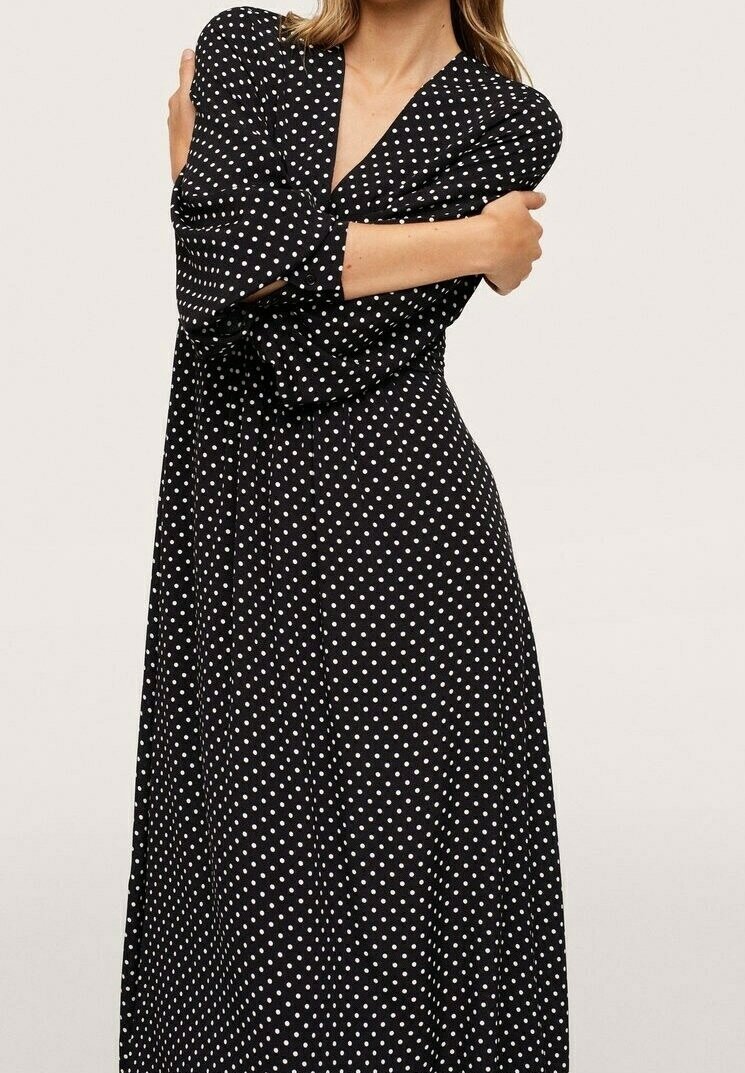 Femme portant une robe noire à manches longues et col en V avec des pois blancs, s’enlaçant devant un fond uni.