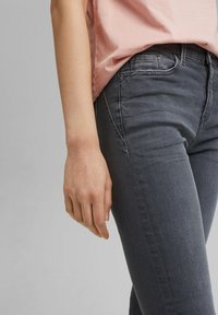 Primer plano de una persona con vaqueros gris oscuro y una camisa rosa claro, mostrando su mano izquierda descansando a su lado.