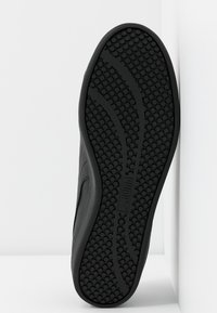 Suela de zapatilla negra con un patrón de agarre texturizado, que presenta puntos en relieve y líneas curvadas, fabricada con materiales de goma y sintéticos.