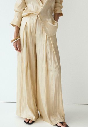 Personne portant un pantalon satiné beige ample et plissé ainsi qu'une chemise boutonnée assortie, accessoirisée avec des bracelets en bois et des sandales noires.