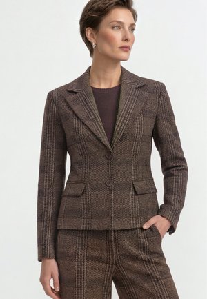 Woman wearing a brown checkered blazer and matching trousers with one hand in pocket, looking to the side against a plain background.