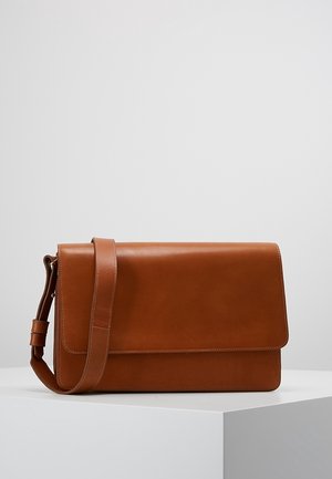 Sac bandoulière en cuir marron avec fermeture à rabat et sangle réglable, posé sur un socle blanc sur un fond uni.