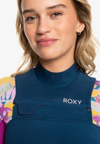 Top de neopreno con un cuerpo azul marino, mangas florales coloridas y el logo de Roxy. Presenta una textura suave y detalles de tirantes ajustables.