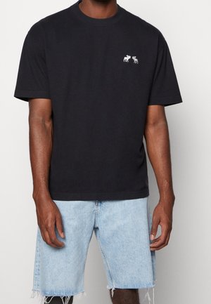 Mand iført sort t-shirt med korte ærmer med lille hvid elg-logo på brystet og lyseblå denimshorts med frynset kant.