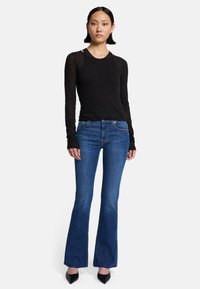 Czarny top z długim rękawem w połączeniu z niebieskimi dżinsami typu dzwony. Top ma przezroczyste, teksturowane rękawy; dżinsy mają klasyczny krój i postrzępiony dół. Całość dopełniają czarne szpilki.