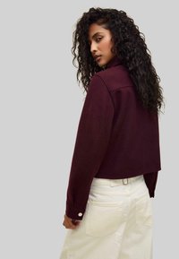 Burgundy cropped jacket with a collar, long sleeves, and buttoned cuffs. Worn over white high-waisted pants, showing a smooth texture.