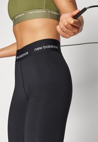 Juodos tamprės su aukštu liemeniu, ant kurių yra pilkos spalvos "New Balance" ženklas. Su derinamu žaliu sportiniu liemenėliu.