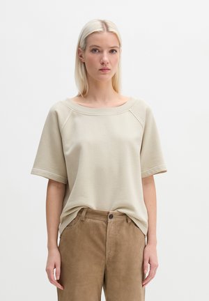 Femme blonde portant un haut beige à manches courtes rentré dans un pantalon taille haute marron clair, debout devant un fond blanc uni.