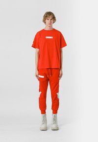 Camiseta roja de manga corta con una etiqueta rectangular blanca. Combinada con pantalones jogger rojos que tienen acentos bordados en blanco y bolsillos con cremallera.