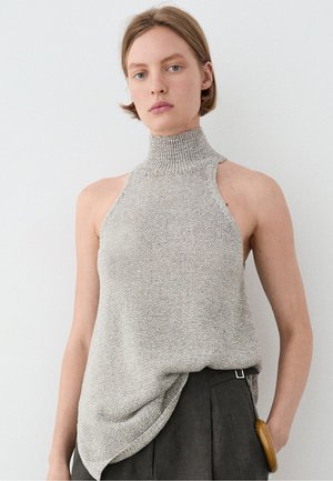 Jeune femme aux cheveux courts porte un haut gris en maille sans manches à col montant et un pantalon foncé, debout devant un fond blanc uni.