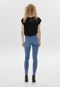 Femme aux cheveux bouclés portant un haut noir à imprimé léopard, un jean skinny bleu et des bottines noires à talons hauts, debout de dos.