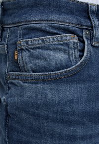 Gros plan d'une poche de jean en denim bleu avec des coutures orange et une petite étiquette noire avec le mot « BOSS » en texte orange.