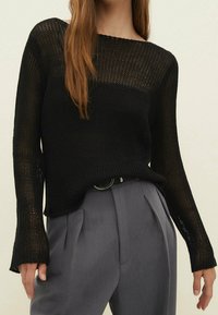 Pull en maille noir à coupe ample, col large et manches longues, associé à un pantalon gris taille haute, avec un tissu texturé.