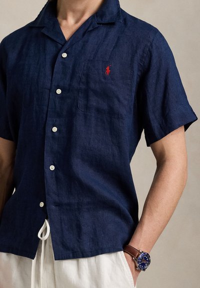 Rövid ujjú, tengerészkék lenvászon ing fehér gombokkal, mellkasi zsebbel és piros logó részlettel. Keskeny szabás, kényelmes illeszkedés.