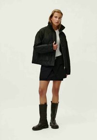 Casaco acolchoado preto com colarinho alto, top branco, saia preta e botas escuras até ao joelho. Corte reto e detalhes de hardware mínimos.