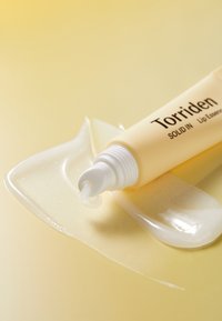 Gul tube af Torriden læbeserum med hvidt låg, der har en glat, blank tekstur, placeret ved siden af et klart gel-lignende stof på en lys baggrund.