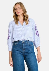Femme portant un jean taille haute bleu et une blouse blanche avec des rayures verticales bleues et des broderies florales violettes sur les manches.