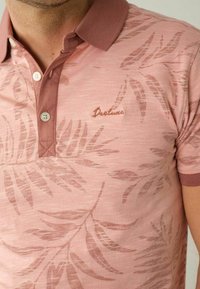 Rosa Polohemd mit einem dezenten Blattmuster, einem kontrastierenden Kragen und einem kleinen gestickten Logo auf der Brust. Es hat eine strukturierte Oberfläche.