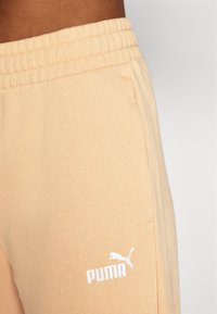 Persiku krāsas sporta bikses, kas izgatavotas no mīksta auduma, ar elastīgu jostu un baltu Puma logo apakšējā daļā.