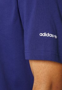 Námořnická modrá bavlněná tričko s kulatým výstřihem; má krátké rukávy a bílý logo "adidas" s trojlístkem na levém rukávu. Hladká textura.