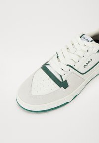 Hvit sneakers med grønne detaljer, med overdel i semsket skinn og lær, perforert tåboks, flate snørebånd og en teksturert gummisåle.