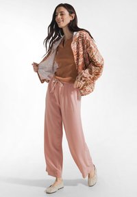 Frau lächelnd, trägt ein braunes Oberteil, eine gemusterte orangefarbene Jacke, rosa Hosen mit Kordelzug und beige Ballerinas, steht vor einem weißen Hintergrund.