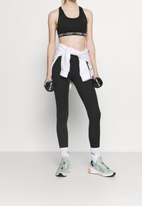 Czarny sportowy stanik z logo na pasku, czarne legginsy, biały sweter związany w talii, szare trampki, trzymający dwa hantle, białe skarpetki.